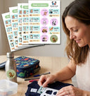 A woman at a desk applies a custom school sticker to a navy blue sweatshirt. In the background, sheets of colorful, cartoon animal-themed name stickers are displayed, along with a matching dinosaur-patterned lunch bag, water bottle, and other school supplies.